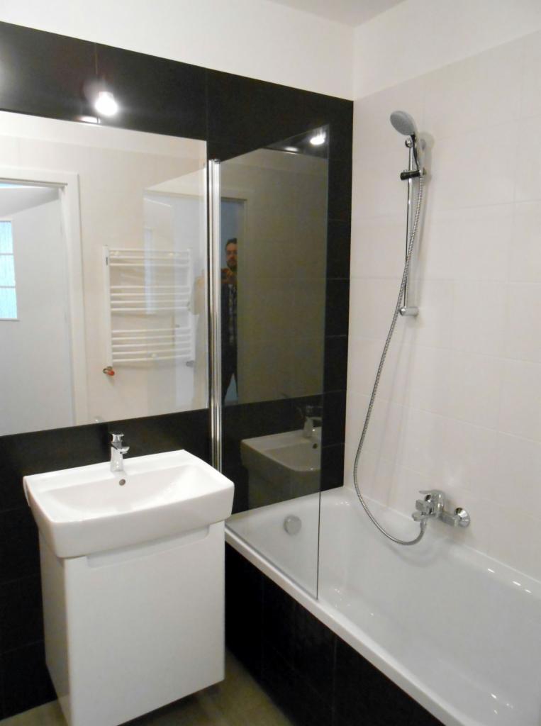 Nowoczesna łazienka z białą umywalką na szafce, wanną z parawanem szklanym i czarnymi akcentami płytek, widok na lustro z odbiciem dodatkowej umywalki w głębi.