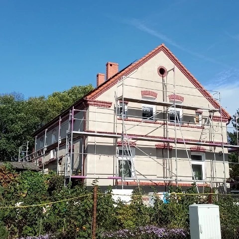 Dwupiętrowy dom w trakcie prac elewacyjnych, widoczne rusztowania, ściany w jasnym kolorze, ceglane detale architektoniczne, słoneczny dzień.