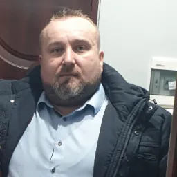 Portret mężczyzny w niebieskiej koszuli i czarnej kurtce na tle ściany i brązowych drzwi.