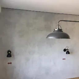 Ściana wykończona szarą, betonową fakturą z widocznymi fugami, zabezpieczona niebieską taśmą malarską przy suficie i krawędzi otworu drzwiowego, z zamontowanymi dwoma czarnymi kinkietami i wiszącą...