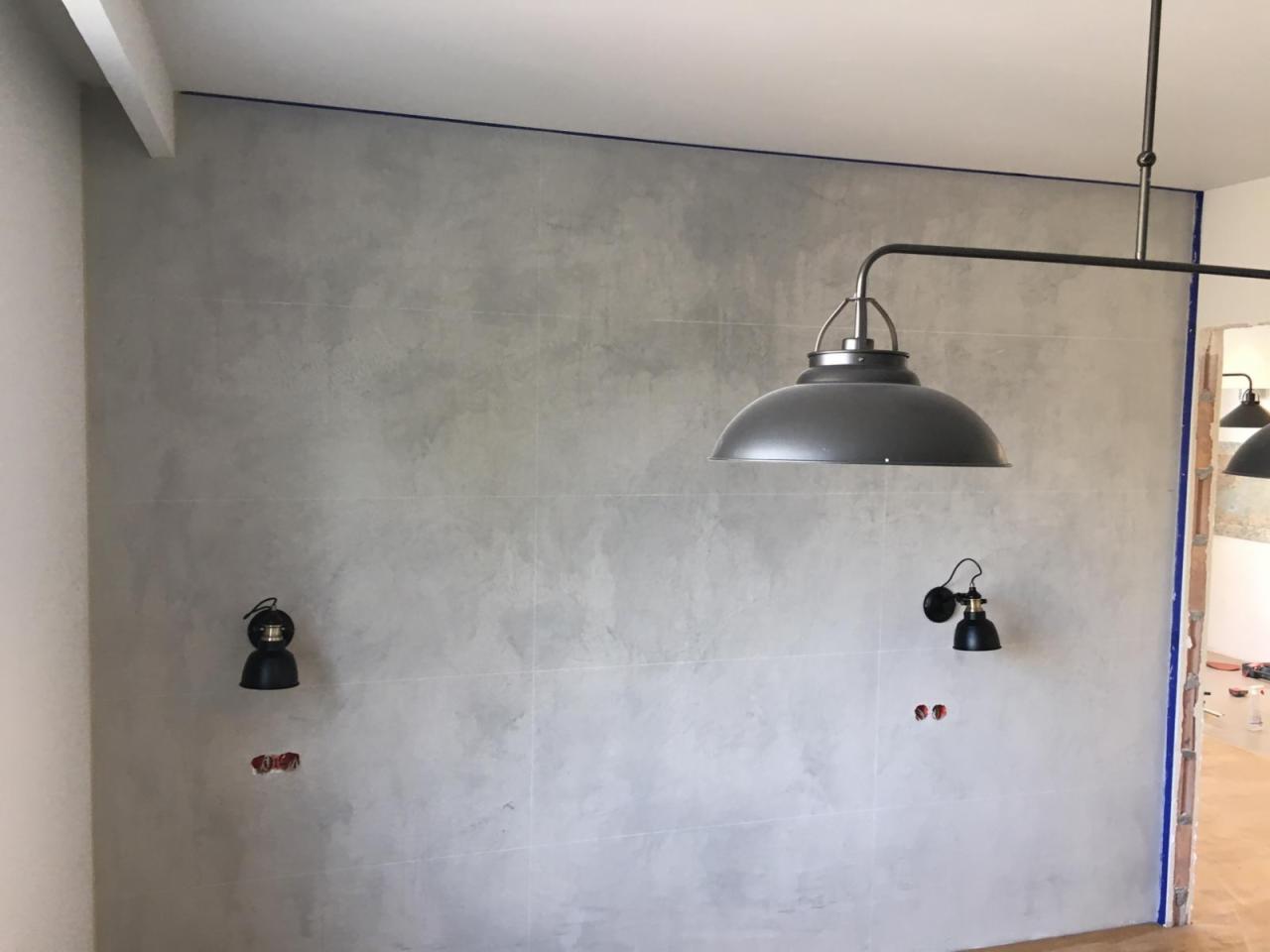 Ściana wykończona szarą, betonową fakturą z widocznymi fugami, zabezpieczona niebieską taśmą malarską przy suficie i krawędzi otworu drzwiowego, z zamontowanymi dwoma czarnymi kinkietami i wiszącą...