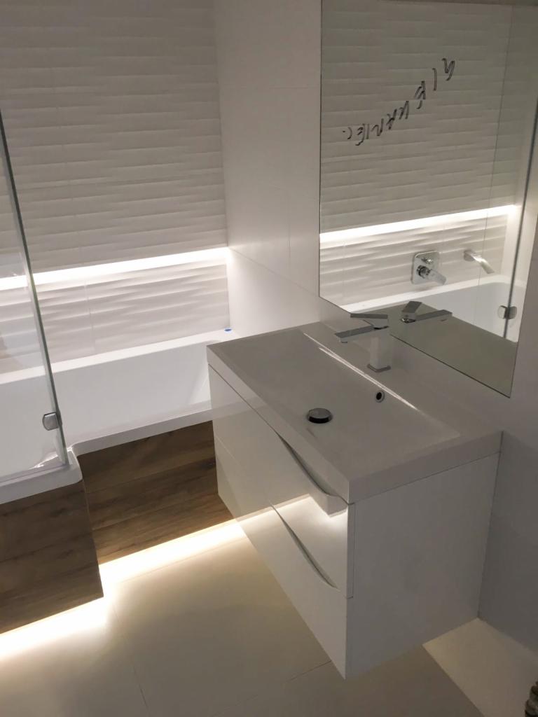 Nowoczesna łazienka w bieli z podświetlanym blatem szafki umywalkowej i lustrem z napisem, widoczna wanna z parawanem nawannowym i drewnopodobna podłoga.