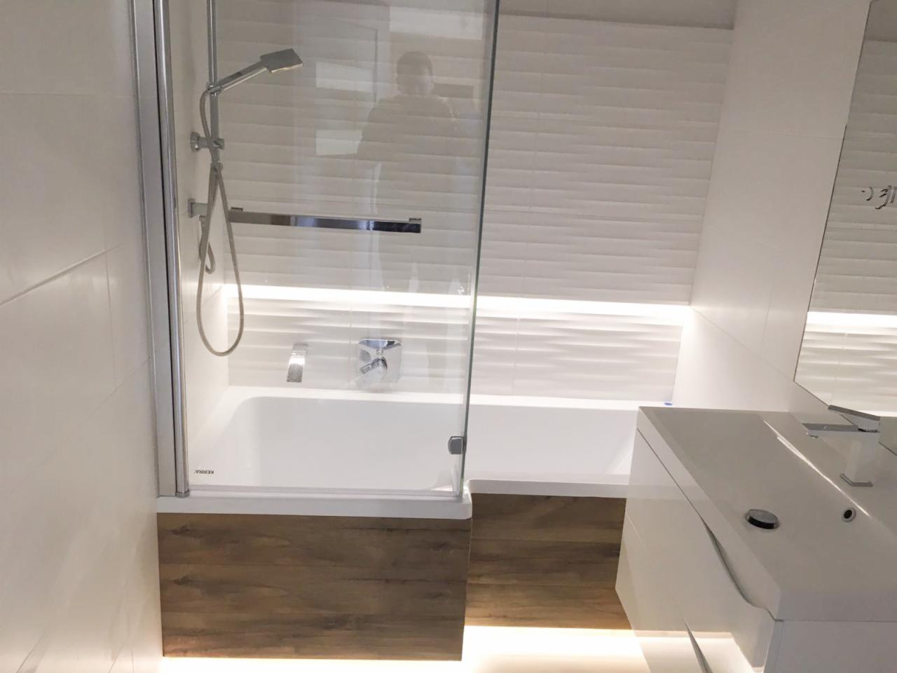 Nowoczesna łazienka z białą wanną obudowaną drewnopodobnymi panelami, szklanym parawanem nawannowym, białą umywalką i podświetleniem LED pod wanną i na ścianie.