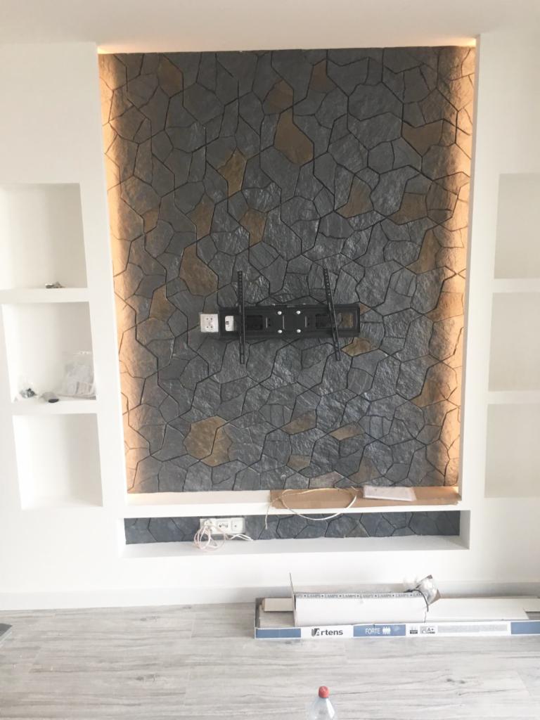 Ściana akcentowa z kamienia dekoracyjnego w salonie, podświetlona od góry, z zamontowanym uchwytem na telewizor i półkami po bokach.