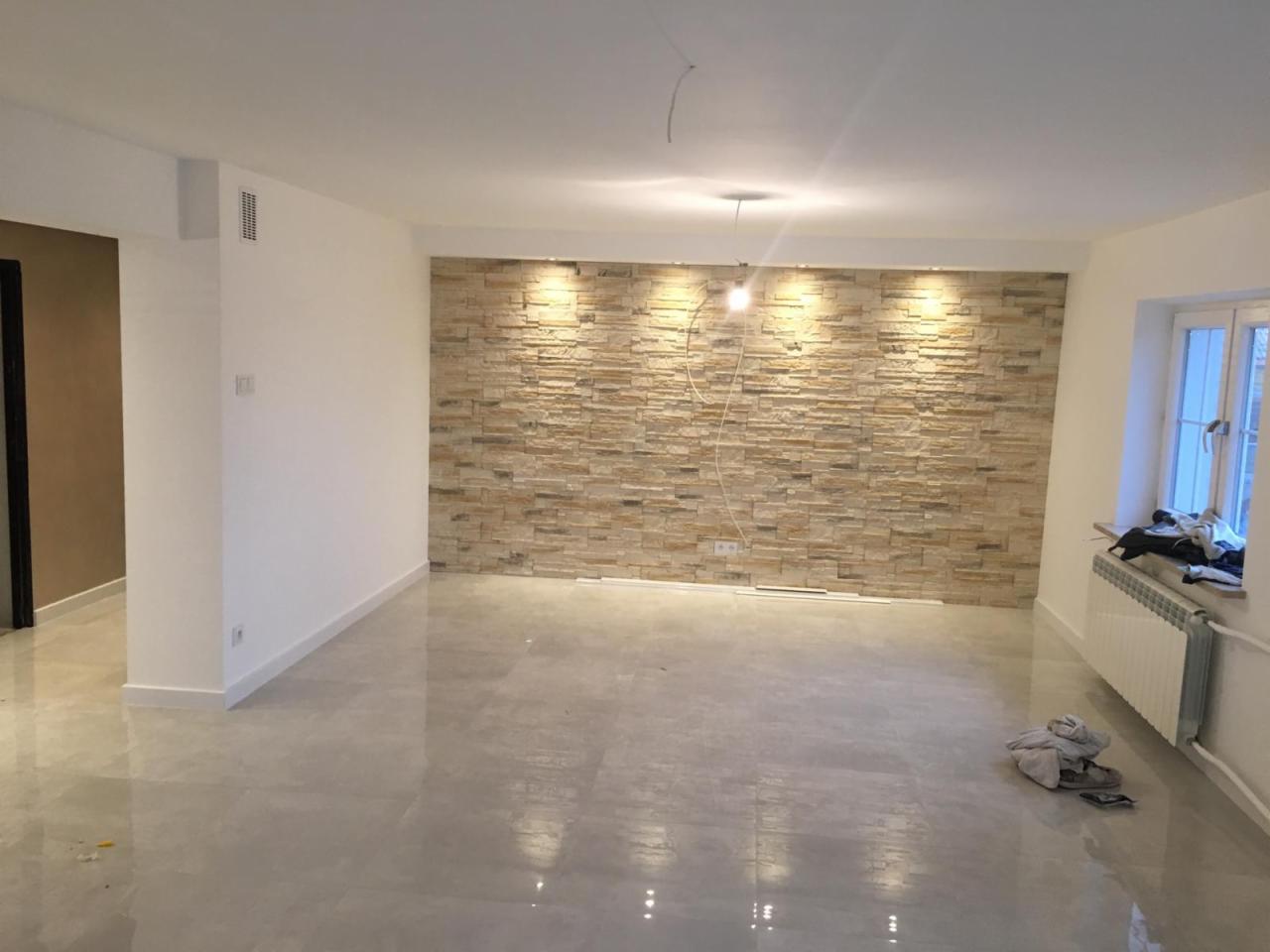 Jasne, przestronne wnętrze po remoncie z akcentem na ścianę wyłożoną dekoracyjnym kamieniem, błyszcząca podłoga z szarych płytek, świeżo pomalowane ściany i widoczny grzejnik pod oknem.