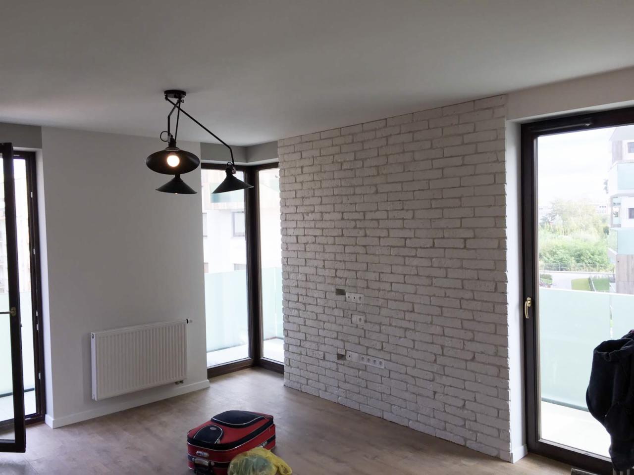 Wnętrze pokoju z białą ceglaną ścianą, drewnopodobną podłogą, czarnymi ramami okiennymi i designerską, trzypunktową lampą sufitową. Widoczne gniazdka elektryczne w ścianie, grzejnik oraz częściowo...