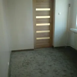 Wnętrze pokoju z szarym dywanem i drewnianymi drzwiami z poziomymi przeszkleniami, widoczny fragment ściany z grzejnikiem.