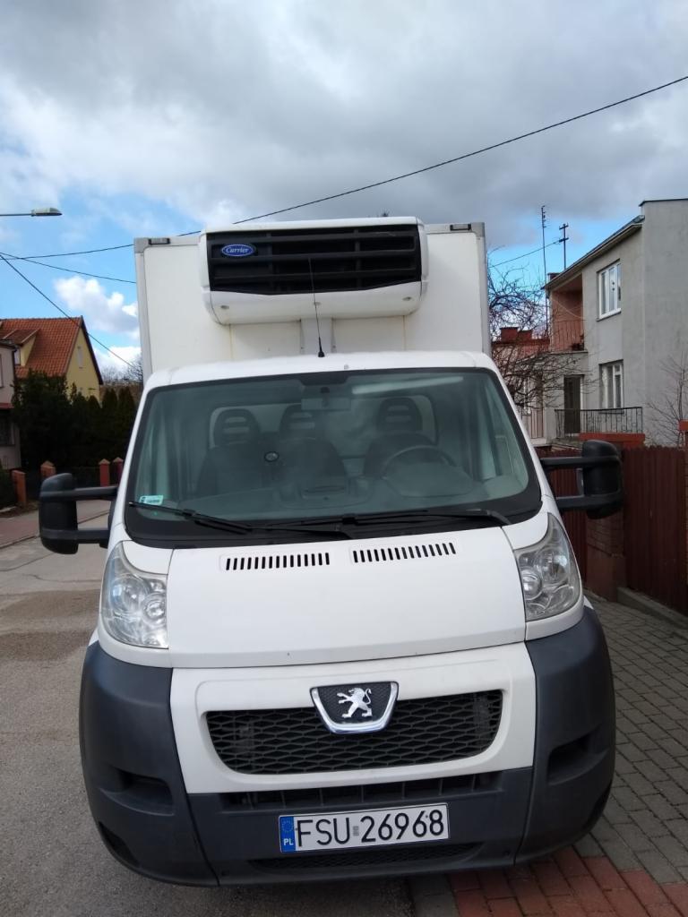 Biały samochód dostawczy z chłodnią Carrier, zaparkowany na ulicy z kostki brukowej, widok z przodu, tablica rejestracyjna FSU 26968.