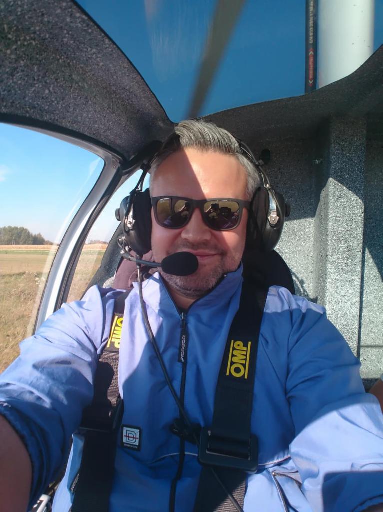 Pilot w kokpicie helikoptera, widoczne słuchawki z mikrofonem, pasy bezpieczeństwa OMP i widok na pole za szybą.