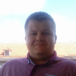 Portret uśmiechniętego mężczyzny w koszuli polo na tle budynku z pomarańczową dachówką i lukarną.