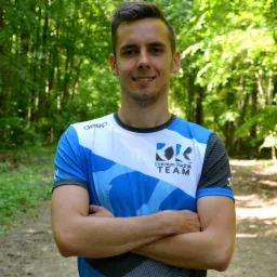 Uśmiechnięty mężczyzna w sportowej koszulce z logo zespołu na tle leśnej ścieżki, ręce skrzyżowane.