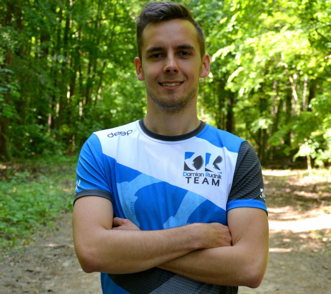 Uśmiechnięty mężczyzna w sportowej koszulce z logo zespołu na tle leśnej ścieżki, ręce skrzyżowane.