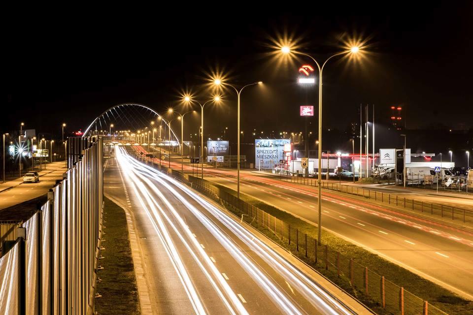 Nocny widok na oświetloną estakadę w Białymstoku z długą ekspozycją, ukazującą smugi świateł samochodów, z widocznymi billboardami i stacją benzynową w tle.