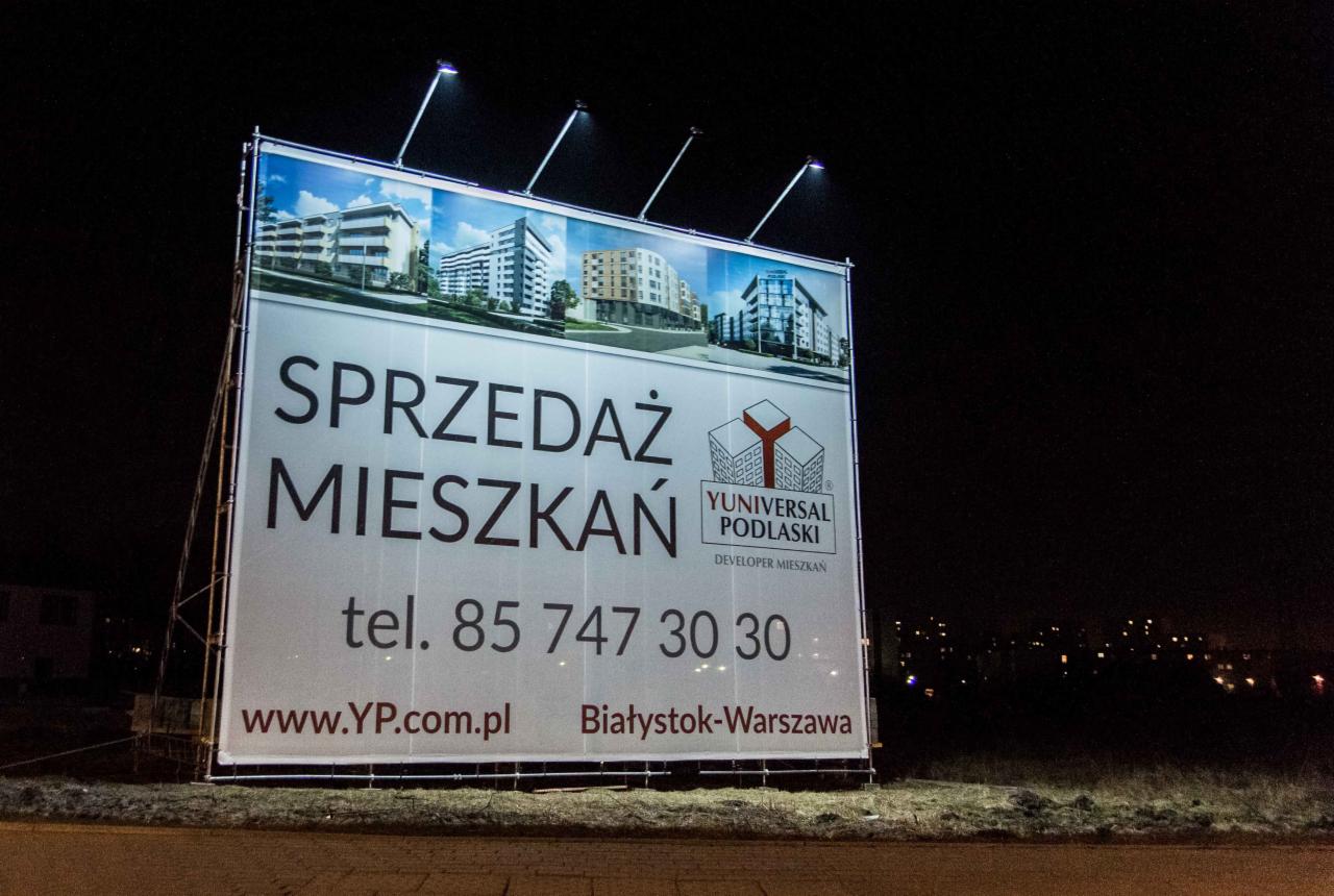 Oświetlony billboard reklamujący sprzedaż mieszkań dewelopera Yuniversal Podlaski, z wizualizacjami budynków mieszkalnych, numerem telefonu i adresem strony internetowej, sfotografowany nocą.