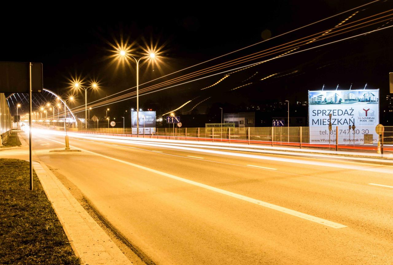 Nocny widok na oświetloną ulicę z billboardem reklamującym sprzedaż mieszkań, widoczne smugi świetlne przejeżdżających samochodów.