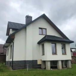 Nowoczesny dom murowany z białymi ścianami, ciemną dachówką i ceglanym kominem na tle pochmurnego nieba, widoczne betonowe podpory fundamentów.