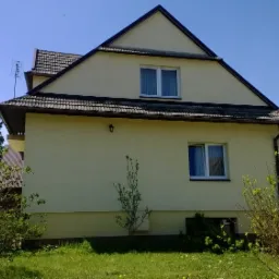 Żółta elewacja dwukondygnacyjnego domu z brązowym dachem, widok z niskiej perspektywy, wiosenne krzewy na pierwszym planie