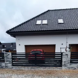 Nowoczesne ogrodzenie z przesuwną bramą w kolorze grafitowym, wykonane z poziomych metalowych profili, osadzone na słupkach z szarej kostki imitującej kamień, na tle dwóch białych domów z ciemnymi...