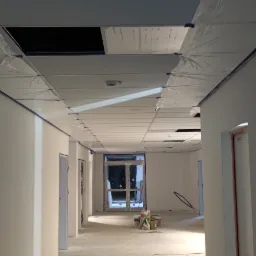 Korytarz w trakcie prac wykończeniowych, ściany pomalowane na biało, sufit zabezpieczony folią malarską, widoczne kable na podłodze, przygotowania do dalszych prac.