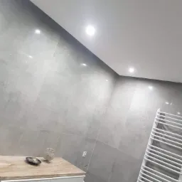 Szare płytki ceramiczne na ścianie w łazience, biała szafka z blatem imitującym marmur, dekoracje na blacie oraz biały grzejnik drabinkowy.