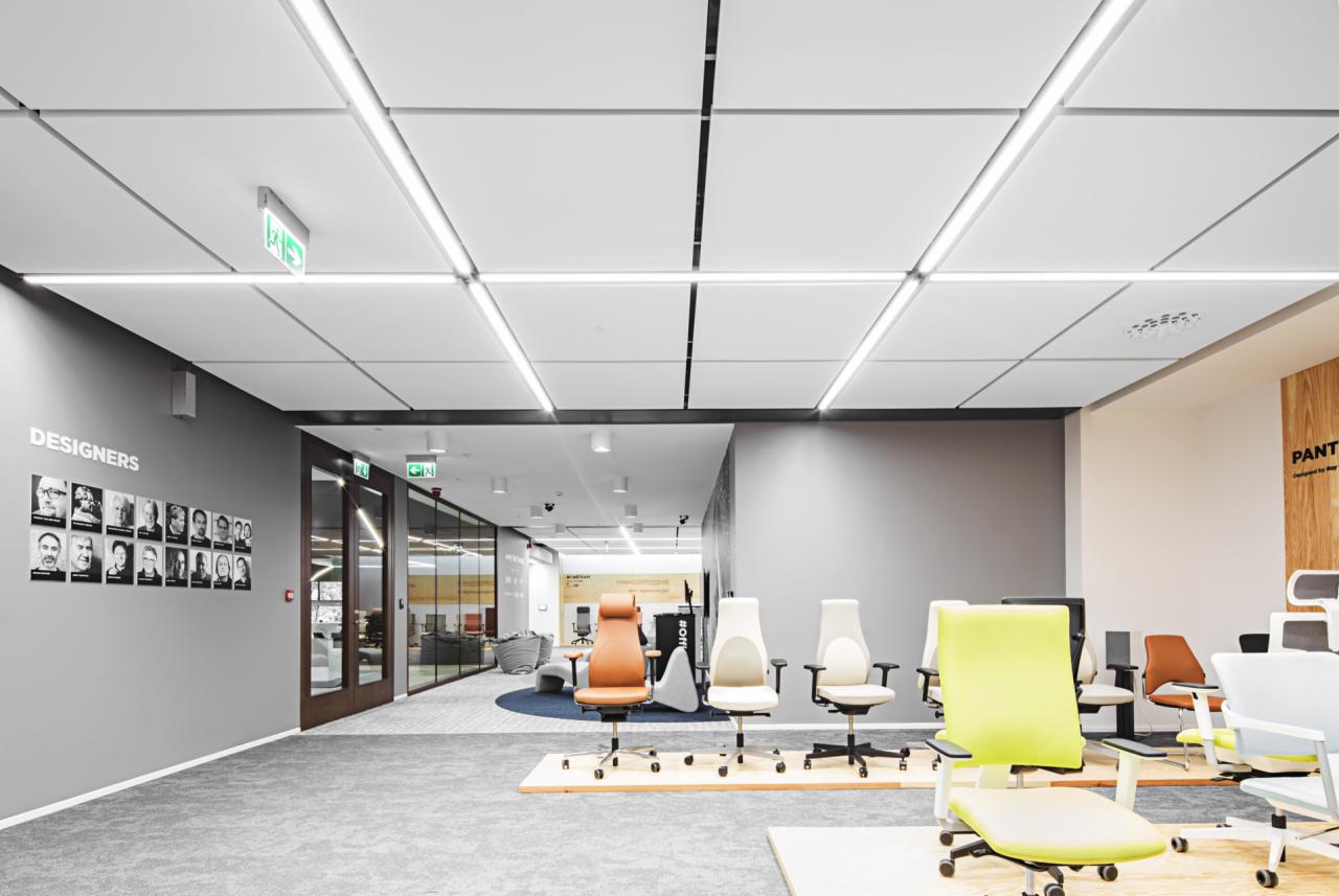 Jasne, nowoczesne biuro z rzędem ergonomicznych krzeseł biurowych w różnych kolorach, podwieszany sufit z liniowym oświetleniem LED, ściana z portretami projektantów.
