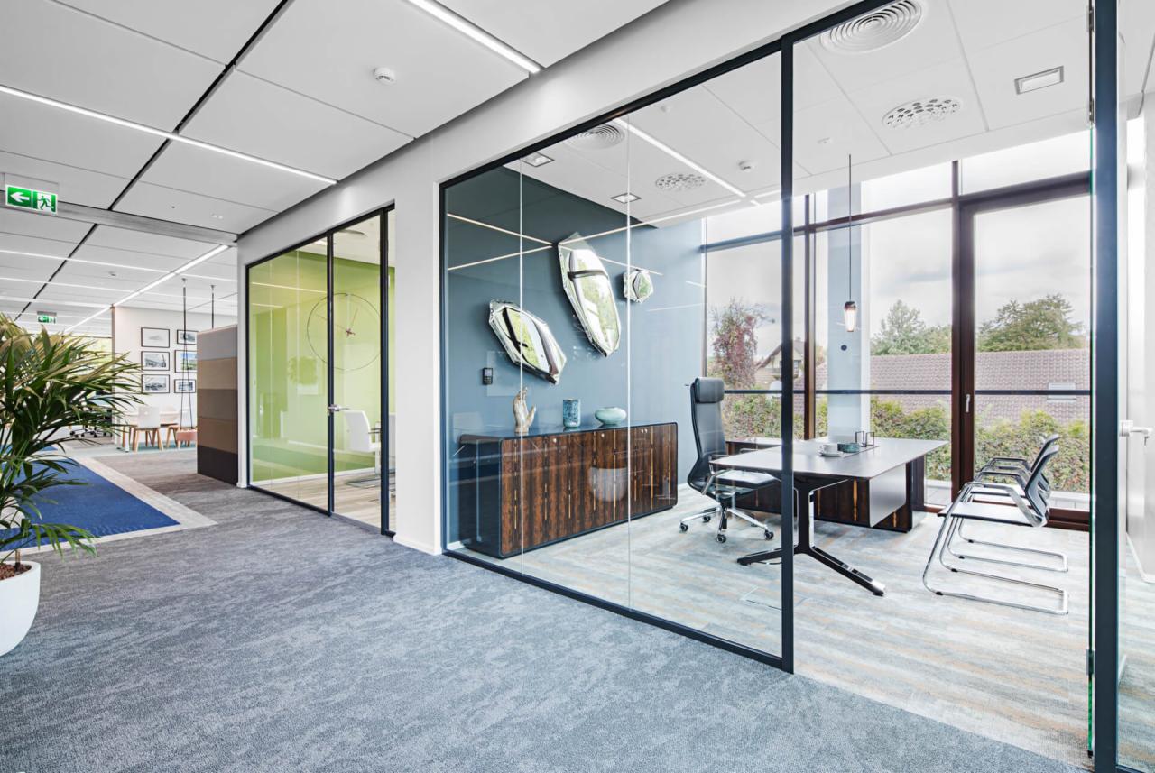 Nowoczesne biuro z przeszklonymi ścianami działowymi, szary dywan, drewniana komoda z dekoracjami i biurko z krzesłami.