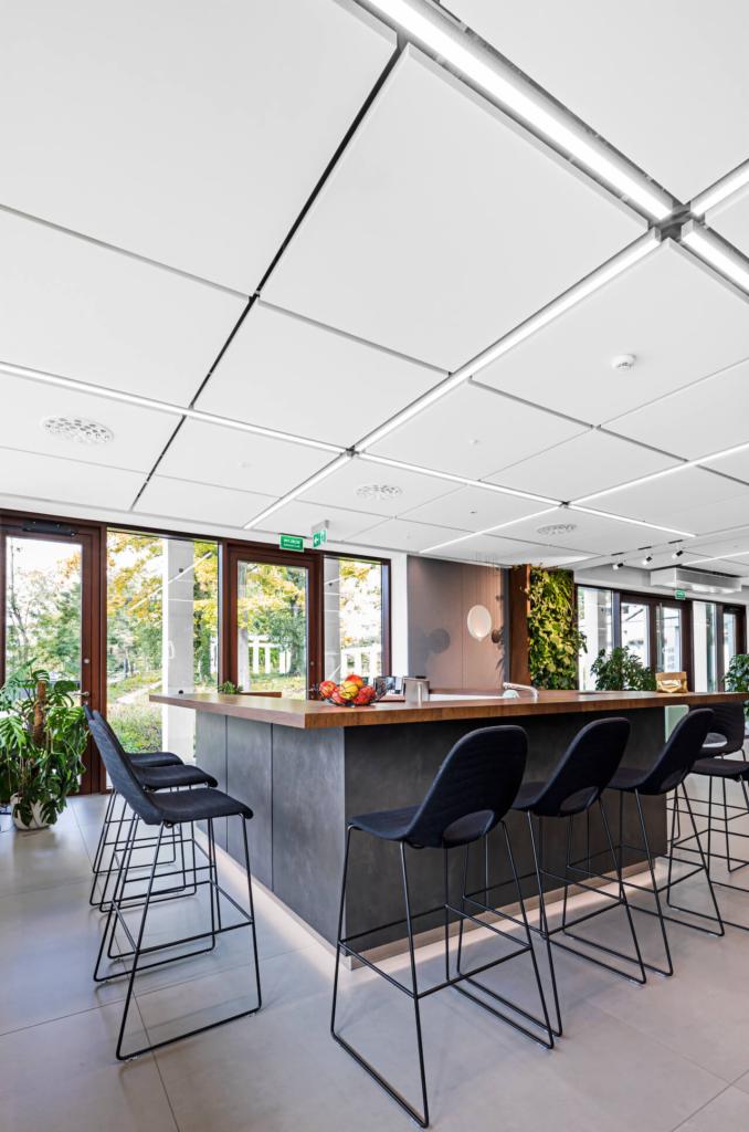Nowoczesna przestrzeń biurowa z wysokim barem, czarnymi krzesłami barowymi, drewnianym blatem, ścianą z zieleni i widokiem na zewnątrz przez duże okna.