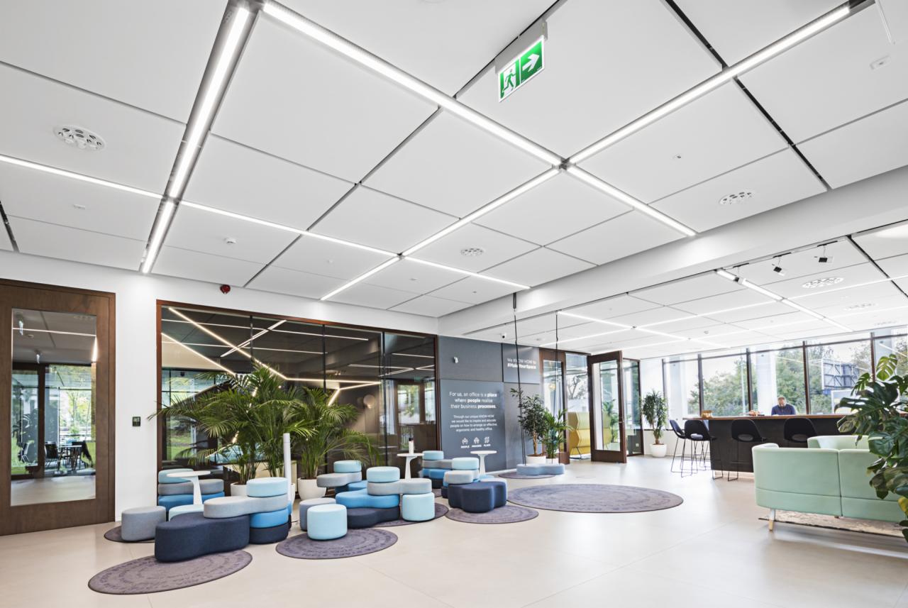 Jasne, nowoczesne wnętrze biurowe z modułowymi siedziskami w odcieniach niebieskiego i szarości, roślinami doniczkowymi i białym sufitem z liniowym oświetleniem LED oraz znakiem ewakuacyjnym.