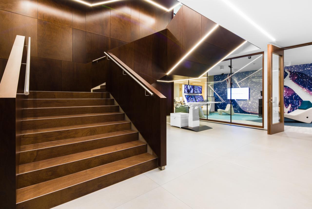 Nowoczesne schody drewniane z metalową poręczą w biurze, oświetlenie LED, widok na przeszkloną salę konferencyjną z futurystycznym wystrojem i ekranami.