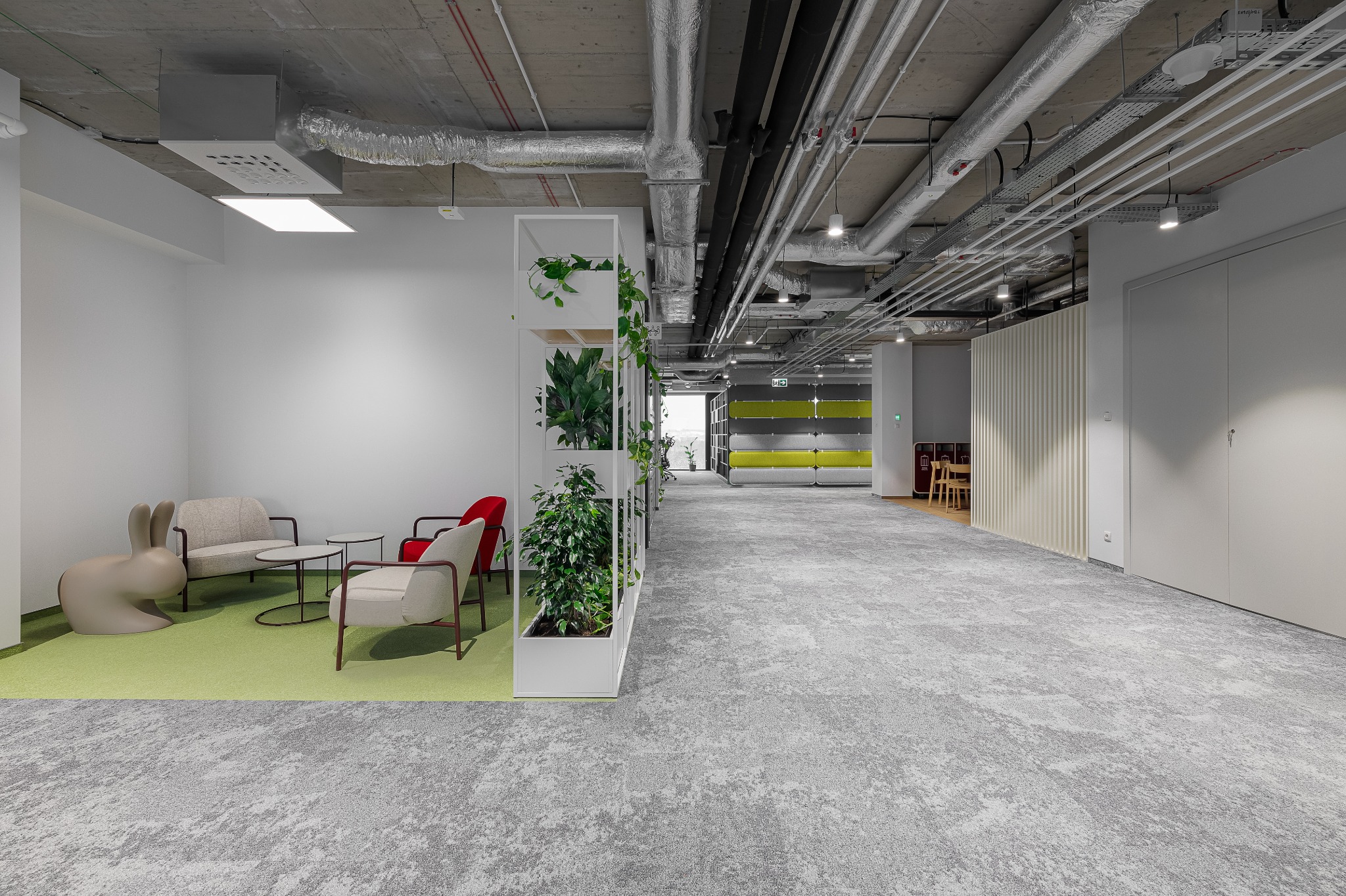 Nowoczesne biuro z otwartą przestrzenią, betonowym sufitem z widocznymi instalacjami, strefą relaksu z beżowymi i czerwonymi fotelami, zielonym dywanem, regałem z roślinami i szarym dywanem...