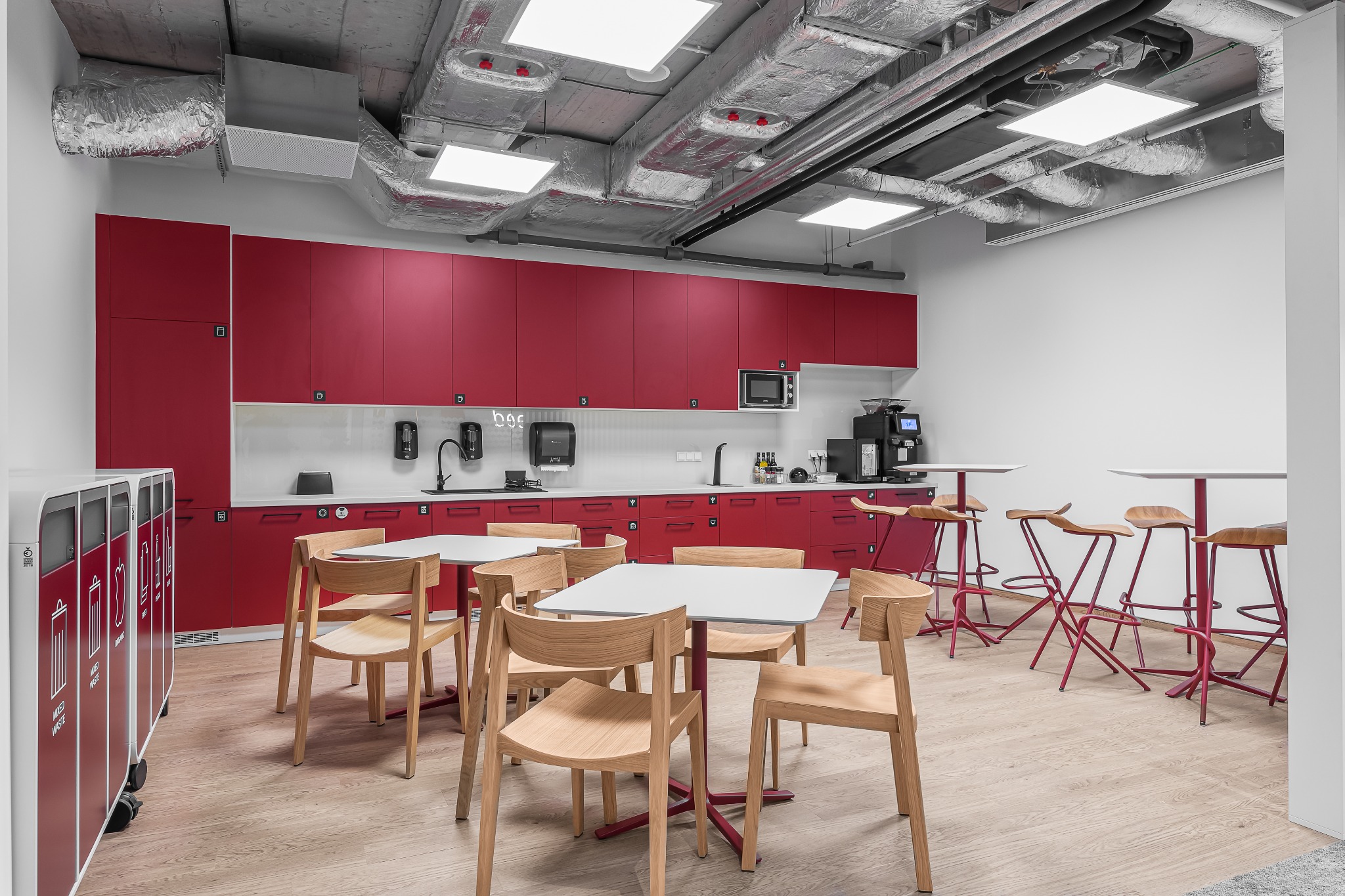 Wnętrze nowoczesnej przestrzeni biurowej z czerwonymi szafkami kuchennymi, drewnianymi krzesłami i stołami na czerwonych nogach, widoczne instalacje wentylacyjne pod sufitem, oraz pojemniki...