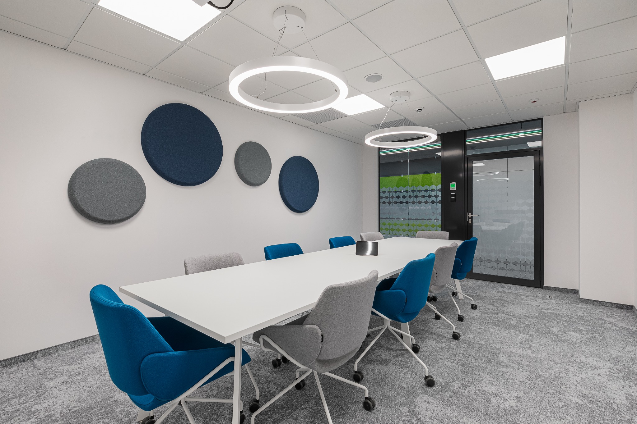 Nowoczesna sala konferencyjna z długim białym stołem, krzesłami w odcieniach szarości i błękitu, okrągłymi lampami LED i panelami akustycznymi na ścianie.