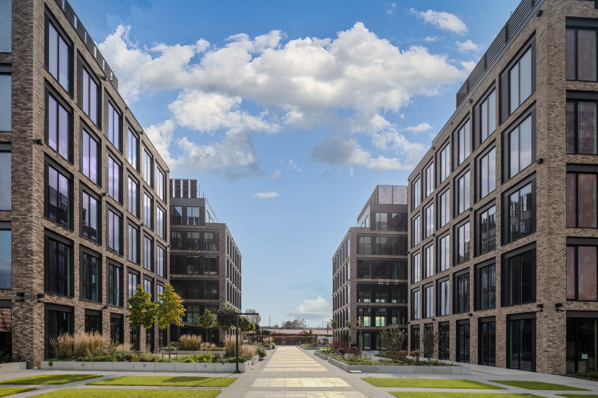 Nowoczesne budynki biurowe z cegły klinkierowej, symetryczne ujęcie z perspektywy chodnika, błękitne niebo z chmurami w tle.