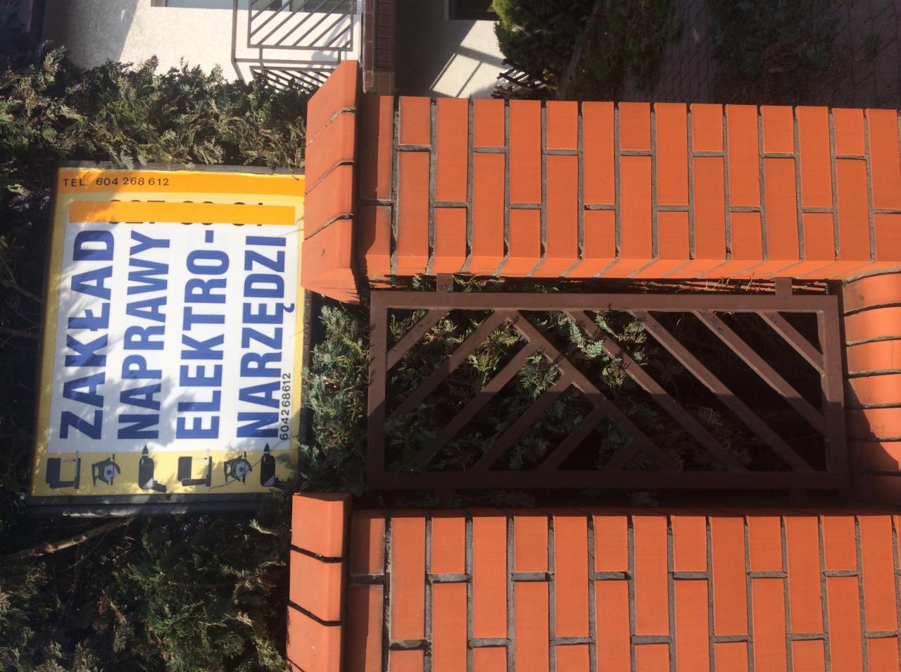 Tablica reklamowa zakładu naprawy elektronarzędzi ustawiona obok ceglanego muru z ozdobną kratą, na tle zieleni.
