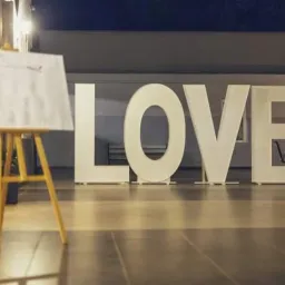 Duże, białe litery układające się w słowo LOVE ustawione na posadzce, w tle rząd krzeseł, z lewej strony sztaluga z białą tablicą i listą gości.