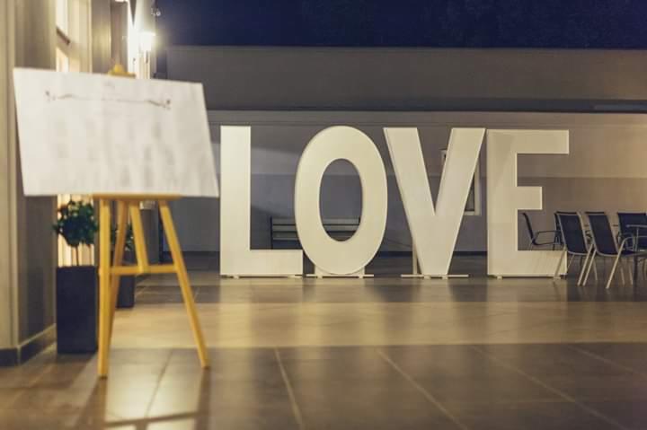 Duże, białe litery układające się w słowo LOVE ustawione na posadzce, w tle rząd krzeseł, z lewej strony sztaluga z białą tablicą i listą gości.