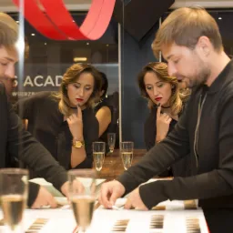 Mężczyzna i kobieta oglądają pr&oacute;bki kolor&oacute;w na stole podczas eventu, w tle widoczne kieliszki z szampanem i logo akademii.