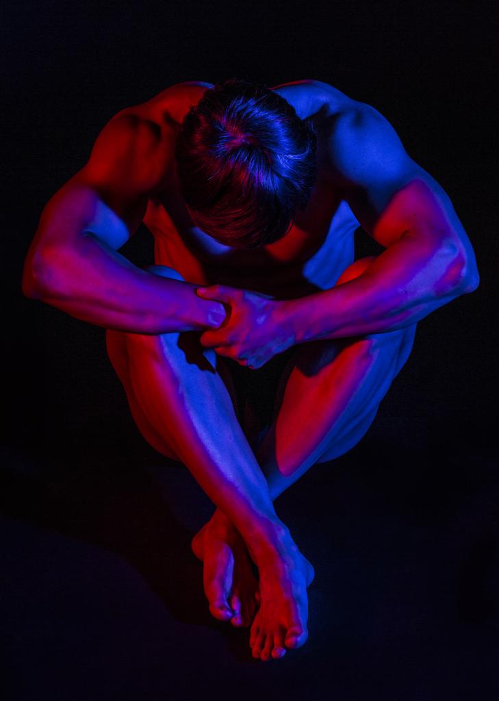 Mężczyzna siedzący ze skrzyżowanymi nogami, głowa pochylona, oświetlony czerwonym i niebieskim światłem na czarnym tle.