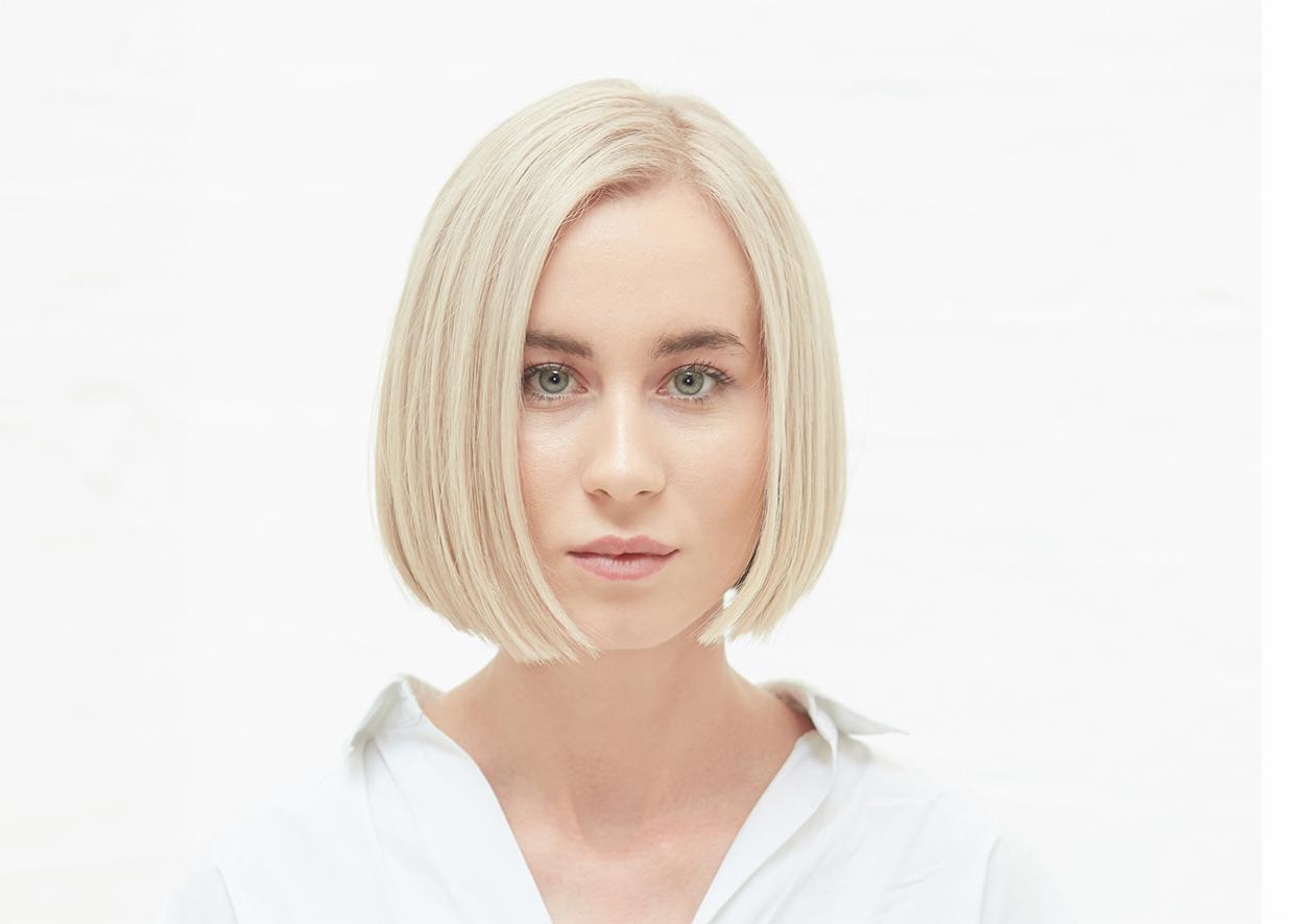 Portret kobiety z krótkimi, platynowymi włosami, ubranej w białą koszulę, na jasnym tle.