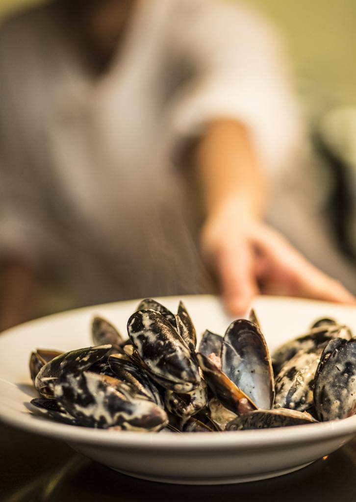 Parujące mule w białym sosie, podawane na białym talerzu, z dłonią w tle.