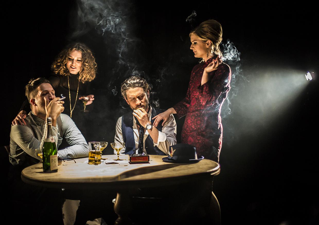 Grupa osób w klimacie retro, siedząca przy okrągłym stole z napojami i papierosami, otoczona dymem i oświetlona punktowym światłem.