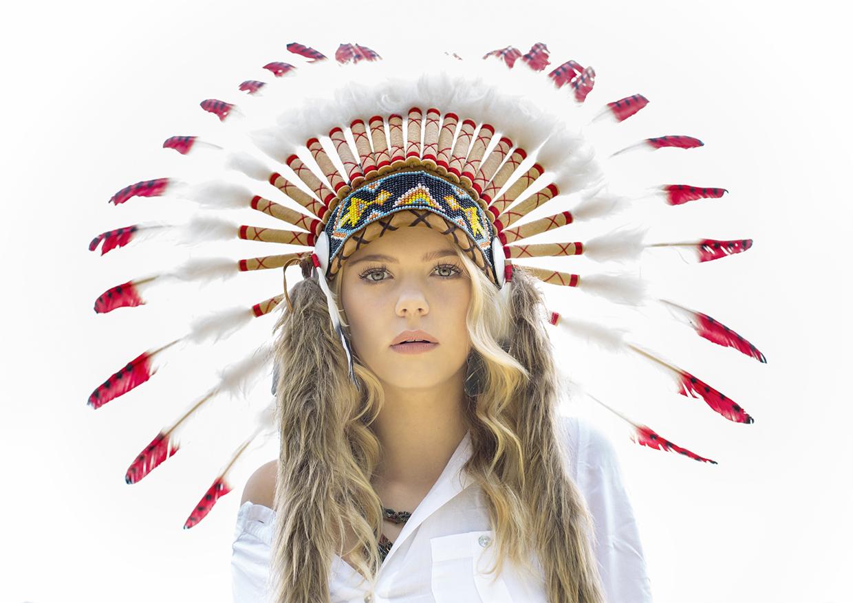 Portret młodej kobiety z blond włosami, ubrana w białą koszulę i tradycyjny indiański pióropusz z białymi i czerwonymi piórami, patrząca prosto w obiektyw na jasnym tle.