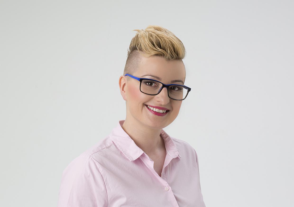 Portret uśmiechniętej kobiety z asymetryczną fryzurą, w okularach z niebieskimi zausznikami i różowej koszuli na jasnym tle.