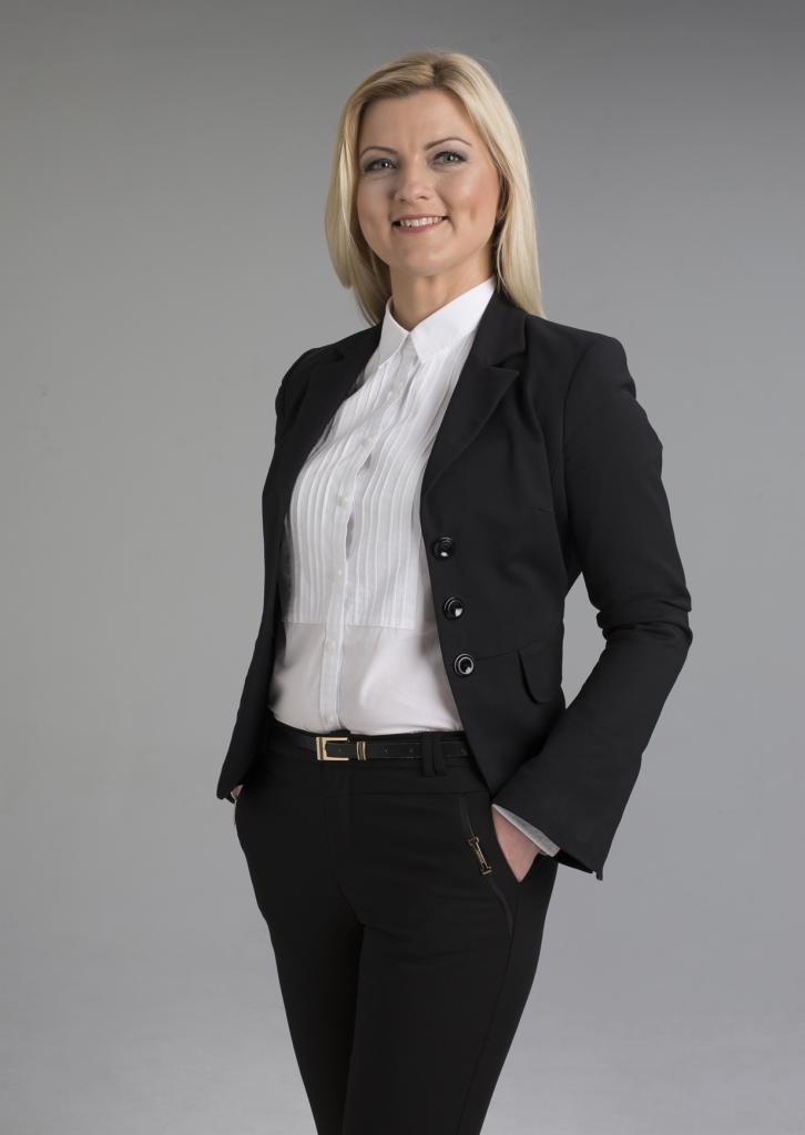 Portret uśmiechniętej blondwłosej kobiety w czarnym garniturze i białej koszuli na neutralnym szarym tle, prezentujący profesjonalny wizerunek.