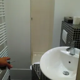 Biała umywalka nablatowa na czarnej mozaice w łazience; w tle kabina prysznicowa, a w kadrze wiertarka trzymana w dłoni w trakcie pracy.