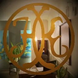 Złoty, ozdobny monogram umieszczony na szklanej powierzchni, przez którą widać fragment biura z roślinami, obrazem i biurkiem.