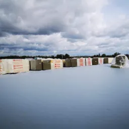 Przygotowane do montażu paczki wełny mineralnej Rockwool ułożone w rzędach na płaskim, szarym dachu, częściowo zabezpieczone folią, na tle pochmurnego nieba.