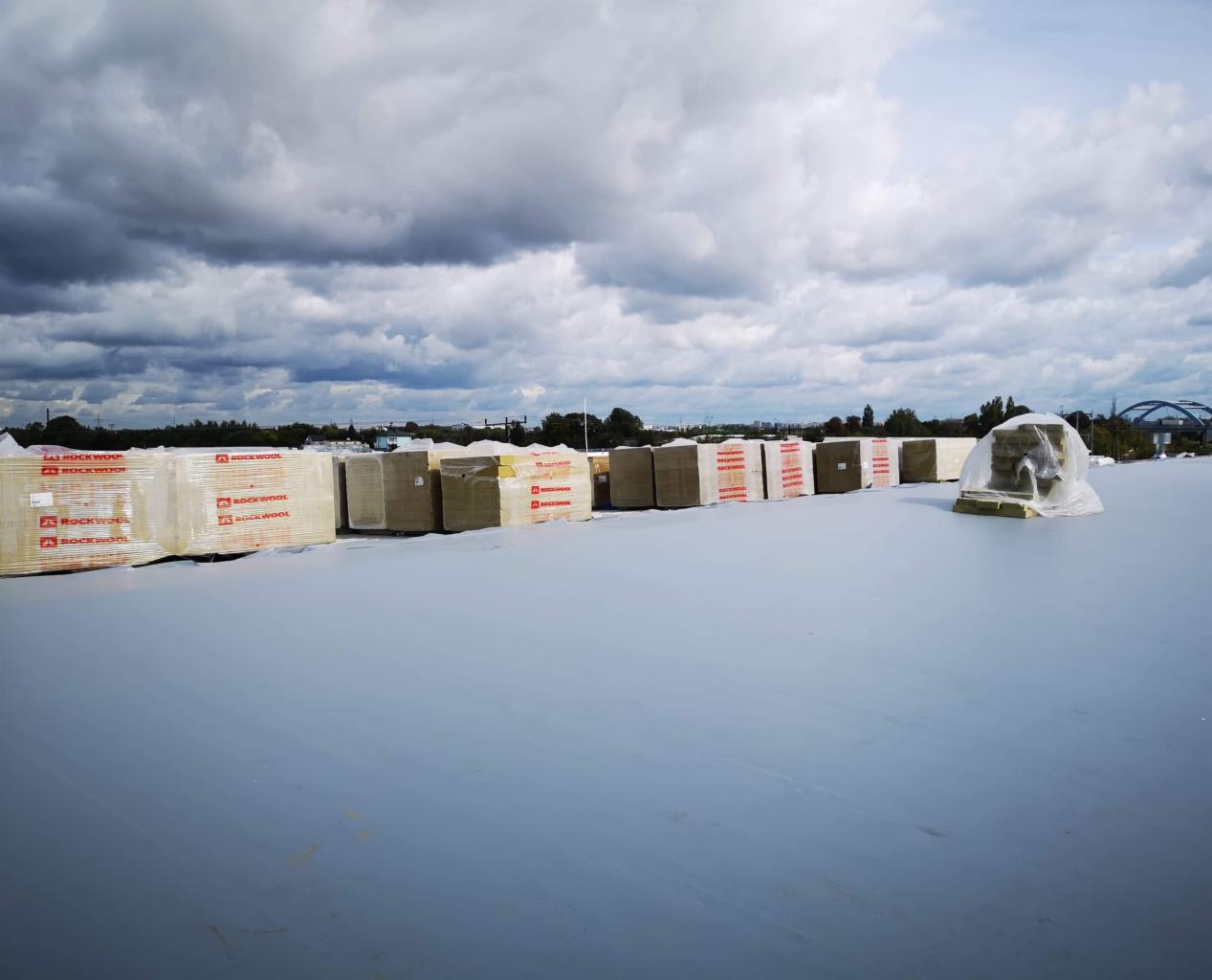 Przygotowane do montażu paczki wełny mineralnej Rockwool ułożone w rzędach na płaskim, szarym dachu, częściowo zabezpieczone folią, na tle pochmurnego nieba.