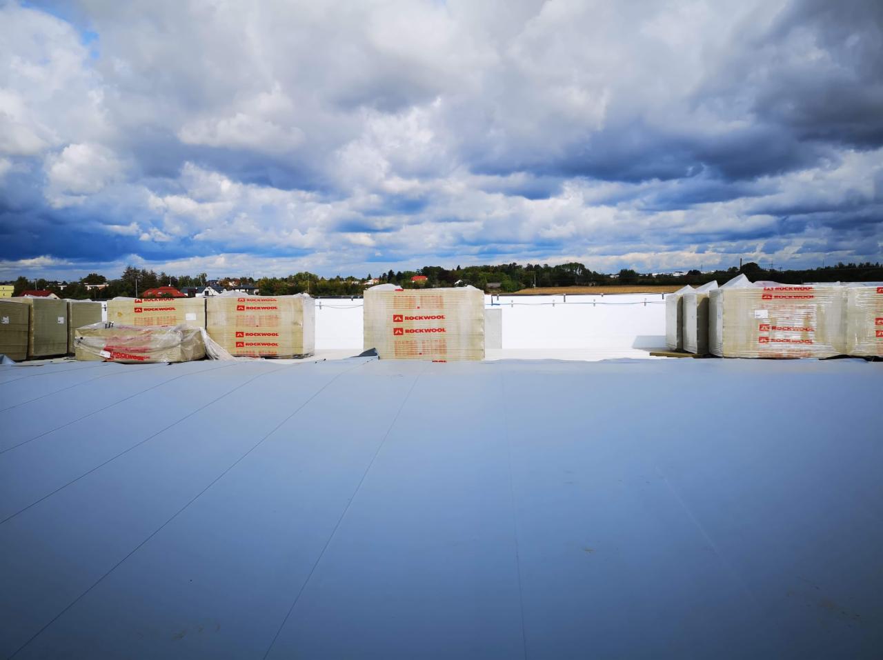 Pakiety izolacji Rockwool na białej, płaskiej powierzchni dachu, z widokiem na oddalony krajobraz pod zachmurzonym niebem.