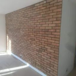 Ściana wykończona cegłą klinkierową w pomieszczeniu, widoczne gniazdka elektryczne i biała listwa przypodłogowa.